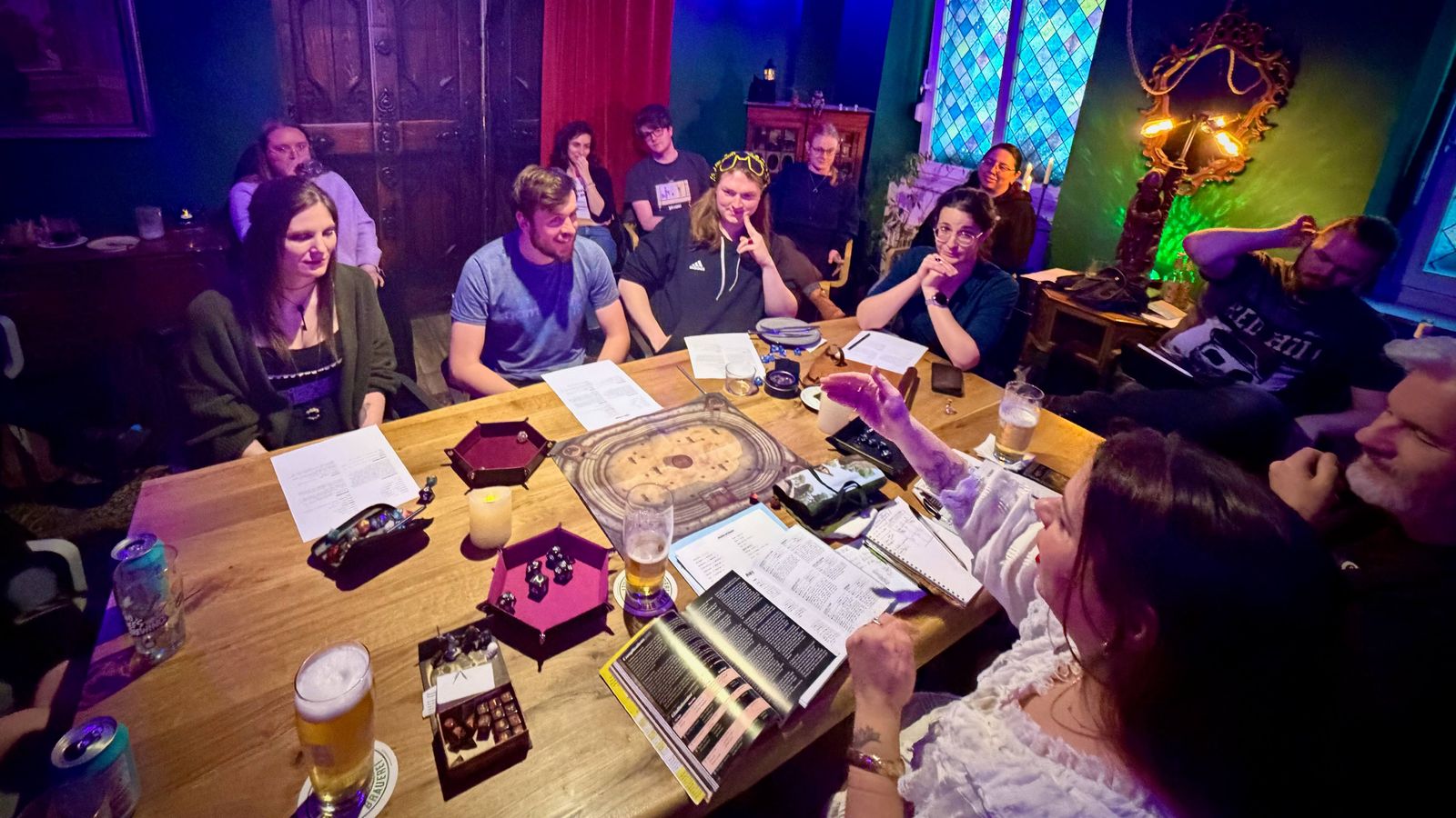 A shot of the RPG room at Tales Untold with Kara as GM gesticulating, and many players and audience members watching and smiling.